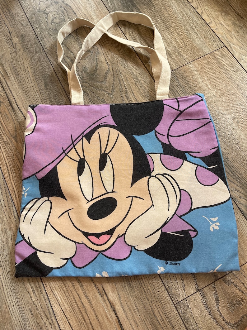May include: A light blue canvas tote bag featuring a purple and white Minnie Mouse design. The bag has white canvas handles and the text "&copy; Disney" is printed on the bottom.