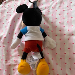 May include: A Mickey Mouse plush keychain. The plush figure has a black head with large ears, a white shirt, blue sleeves, red shorts, yellow shoes, and white gloves. The keychain has a metal ring and a blue fabric loop.