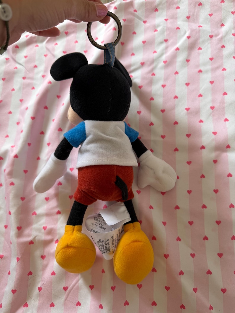 May include: A Mickey Mouse plush keychain. The plush figure has a black head with large ears, a white shirt, blue sleeves, red shorts, yellow shoes, and white gloves. The keychain has a metal ring and a blue fabric loop.