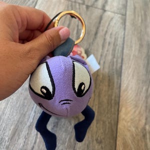 May include: A plush keychain featuring a purple character with large white eyes, black accents, and dark blue legs. The keychain has a gold-colored ring and a small fabric tag.