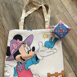 May include: A white tote bag featuring a Minnie Mouse graphic in pink, purple, blue, and black. A small, square pin cushion with a colorful geometric pattern rests on the bag. The bag has beige handles.