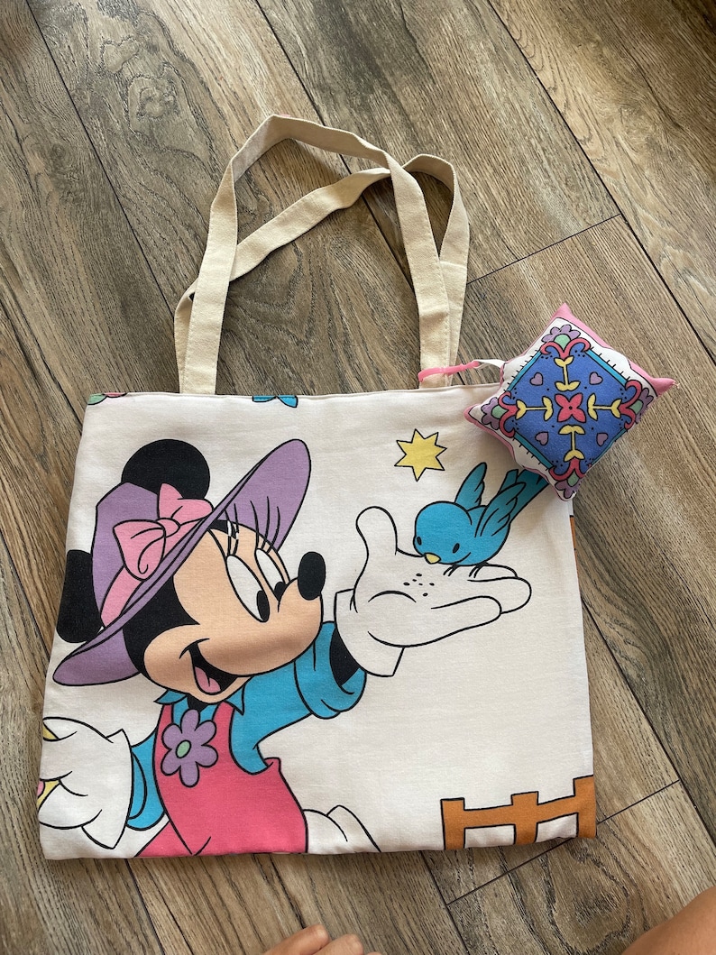 May include: A white tote bag featuring a Minnie Mouse graphic in pink, purple, blue, and black. A small, square pin cushion with a colorful geometric pattern rests on the bag. The bag has beige handles.