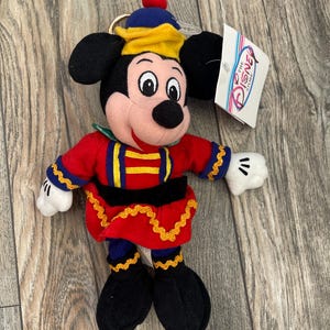 May include: A Mickey Mouse soft toy dressed in a red and blue uniform with gold detailing. The toy has black ears, a black nose, and white gloves. A Disney Store tag is attached.