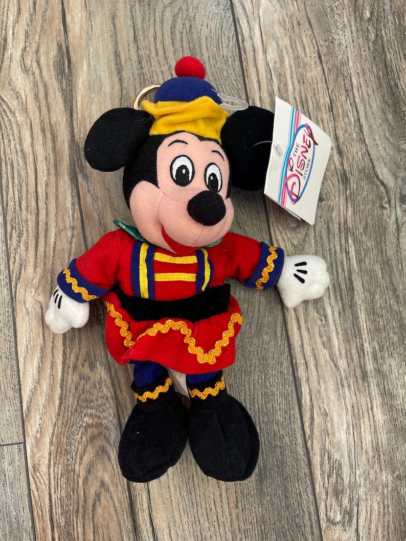 May include: A Mickey Mouse soft toy dressed in a red and blue uniform with gold detailing. The toy has black ears, a black nose, and white gloves. A Disney Store tag is attached.