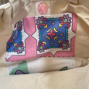 May include: Cream-colored tote bag with a patterned pocket. The pocket features a pink background with blue, yellow, and purple floral designs. The bag has sturdy handles and a tag that reads "The Lion & The Bee".