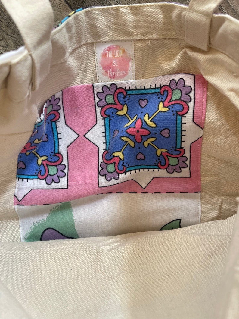 May include: Cream-colored tote bag with a patterned pocket. The pocket features a pink background with blue, yellow, and purple floral designs. The bag has sturdy handles and a tag that reads "The Lion & The Bee".