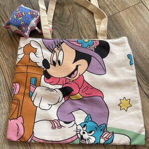 May include: A white tote bag with a Minnie Mouse graphic, featuring a cartoon Minnie in a pink jacket, purple pants, and a purple hat. The bag has cream-colored handles and a small, square, patterned pouch.