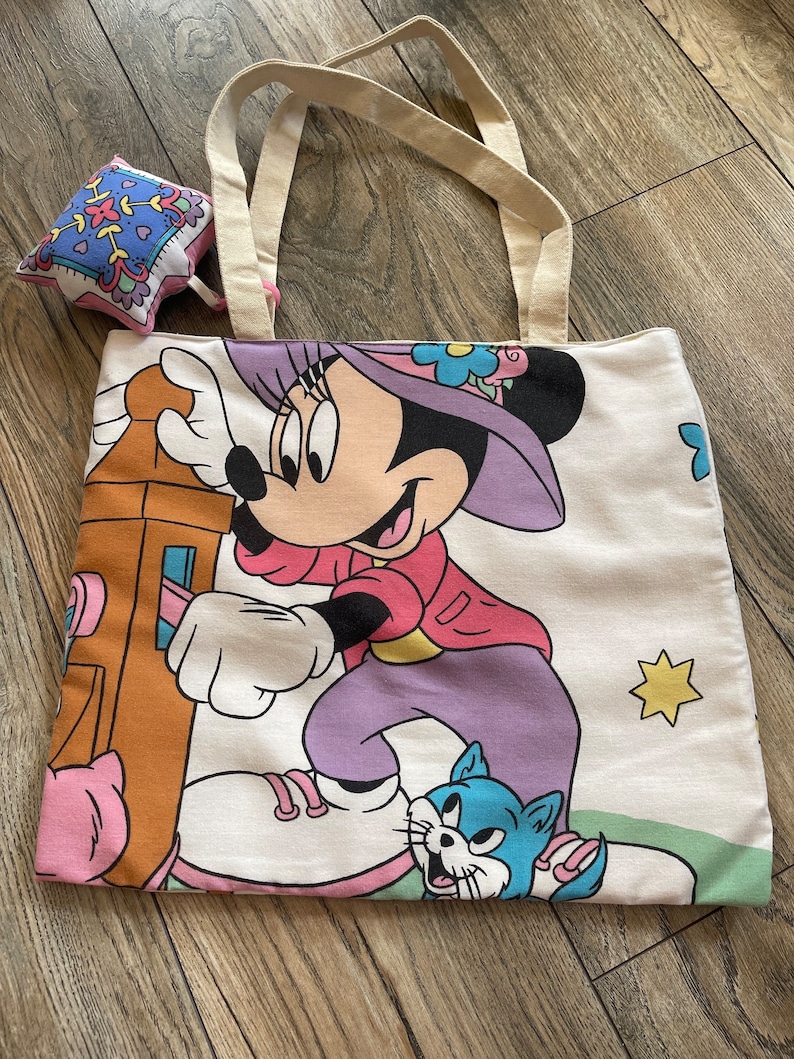 May include: A white tote bag with a Minnie Mouse graphic, featuring a cartoon Minnie in a pink jacket, purple pants, and a purple hat. The bag has cream-colored handles and a small, square, patterned pouch.