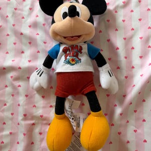 May include: A Mickey Mouse plush toy with black ears, a tan face, and a blue and white shirt with the text "Pixar Pier". The toy has red shorts, yellow shoes, and white gloves. A metal ring is attached to the top of the toy.
