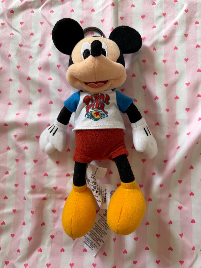 May include: A Mickey Mouse plush toy with black ears, a tan face, and a blue and white shirt with the text "Pixar Pier". The toy has red shorts, yellow shoes, and white gloves. A metal ring is attached to the top of the toy.