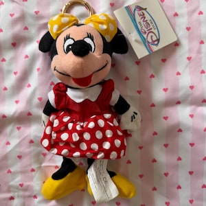 May include: Minnie Mouse plush keychain. The plush toy features a red dress with white polka dots, yellow shoes, and a yellow bow with white polka dots. The keychain has a gold-colored ring and a Disney Store tag.