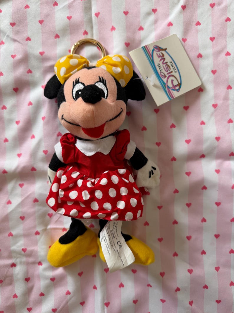 May include: Minnie Mouse plush keychain. The plush toy features a red dress with white polka dots, yellow shoes, and a yellow bow with white polka dots. The keychain has a gold-colored ring and a Disney Store tag.