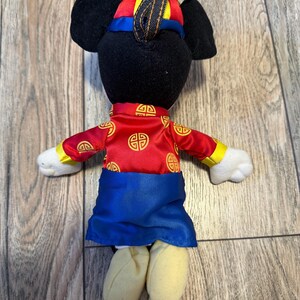 Pu&ograve; includere: Un peluche a forma di topo che indossa un abito rosso e blu con dettagli dorati. Il topo ha orecchie nere e un cappello abbinato. L'abito presenta un design tradizionale. Il giocattolo &egrave; su una superficie di legno.