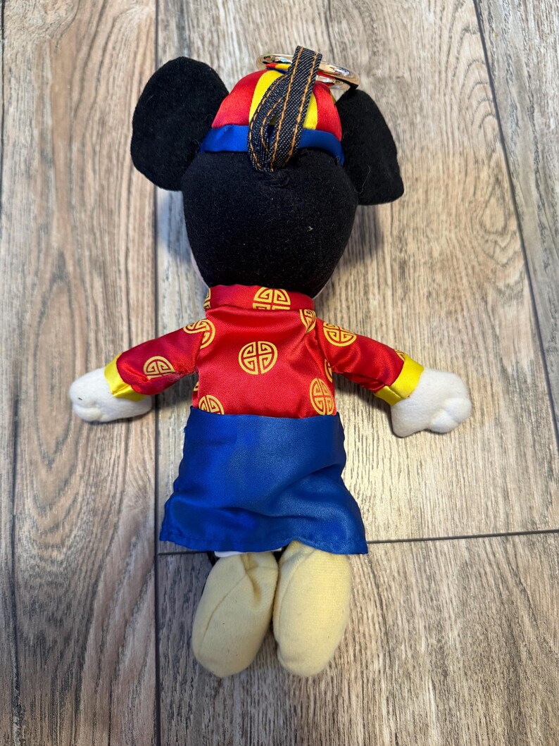 Pu&ograve; includere: Un peluche a forma di topo che indossa un abito rosso e blu con dettagli dorati. Il topo ha orecchie nere e un cappello abbinato. L'abito presenta un design tradizionale. Il giocattolo &egrave; su una superficie di legno.