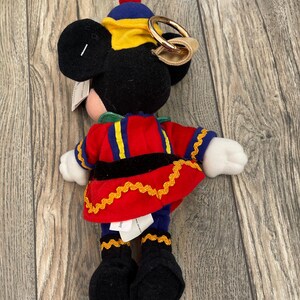 May include: A Mickey Mouse soft toy dressed in a red, blue, and yellow uniform with gold detailing. The toy has black ears, a red bobble on its hat, and a gold-coloured ring attached to its head. The plush is on a wooden surface.