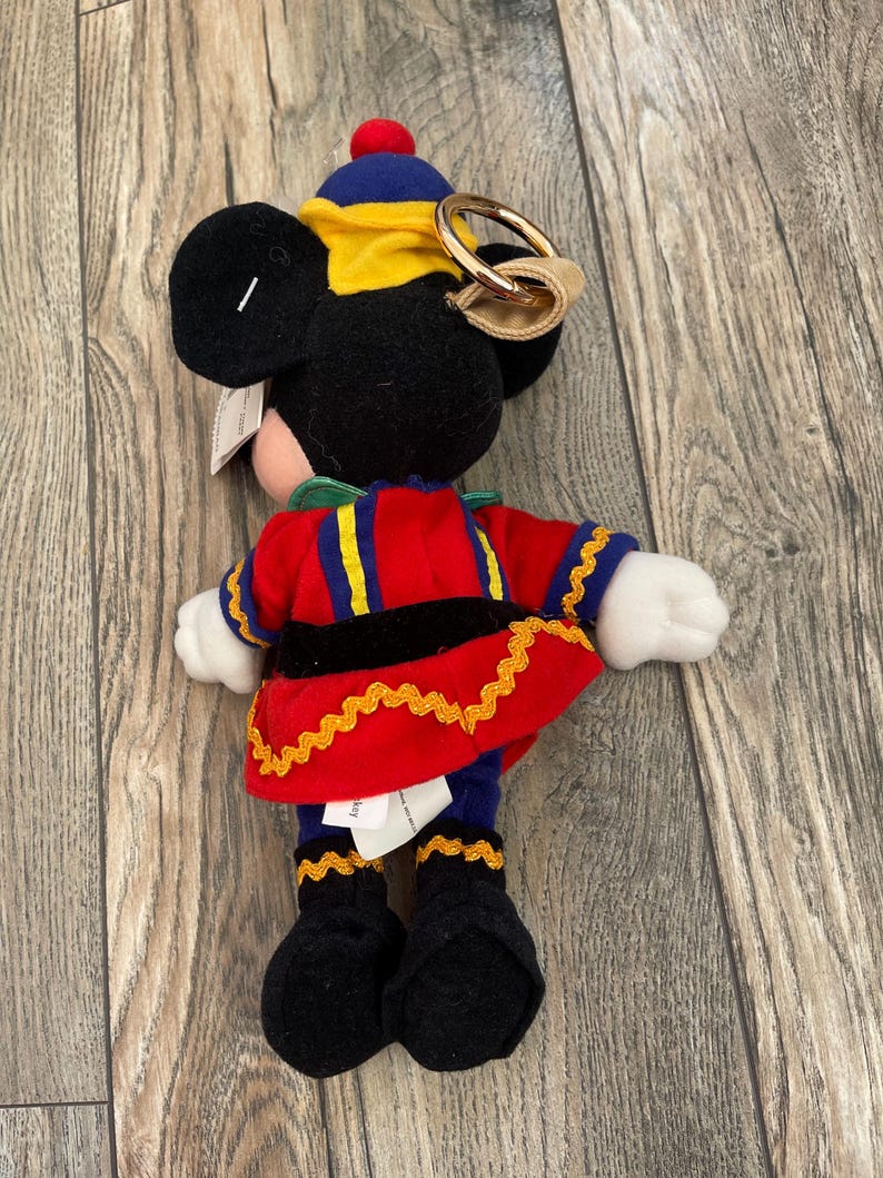 May include: A Mickey Mouse soft toy dressed in a red, blue, and yellow uniform with gold detailing. The toy has black ears, a red bobble on its hat, and a gold-coloured ring attached to its head. The plush is on a wooden surface.