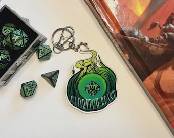 May include: A keychain with a green and yellow Eldritch Blast design, with a silver clasp. Beside the keychain are several dice, including a set of green and black polyhedral dice and a black triangular die. A Player's Handbook is in the background.