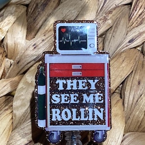 May include: A glittery badge reel with a medical theme. The top features a monitor displaying a heartbeat, and the main body reads "THEY SEE ME ROLLIN" in white letters. The reel has a red and white design with a green oxygen tank.