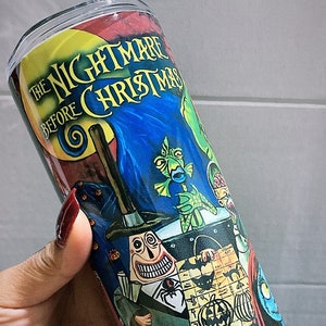 K&ouml;nnte beinhalten: Ein zylindrischer Becher mit einem farbenfrohen Design aus "The Nightmare Before Christmas". Der Becher zeigt Charaktere und Szenen aus dem Film, wobei der Titel deutlich sichtbar ist. Das Design umfasst eine Vielzahl von Farben, darunter Blau, Rot und Gr&uuml;n.
