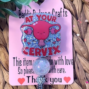 May include: A pink and blue glitter badge reel with a cartoon uterus and the text "At Your Cervix". The reel is attached to a clear crystal bead and a silver retractable badge holder. The background is a woven wicker surface.