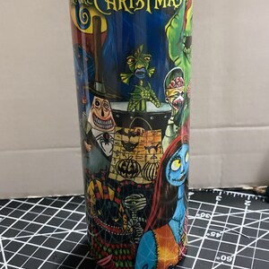 K&ouml;nnte beinhalten: Ein hoher, zylindrischer Becher mit einem farbenfrohen Design aus "The Nightmare Before Christmas". Der Becher zeigt verschiedene Charaktere und Szenen aus dem Film, mit dem Titel in Gelb. Der Hintergrund ist eine Mischung aus dunklen Blau- und Gr&uuml;nt&ouml;nen.