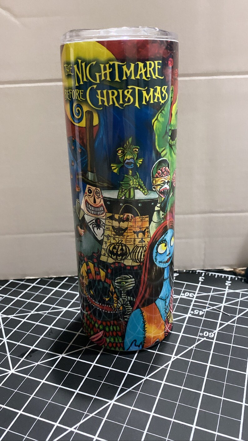 K&ouml;nnte beinhalten: Ein hoher, zylindrischer Becher mit einem farbenfrohen Design aus "The Nightmare Before Christmas". Der Becher zeigt verschiedene Charaktere und Szenen aus dem Film, mit dem Titel in Gelb. Der Hintergrund ist eine Mischung aus dunklen Blau- und Gr&uuml;nt&ouml;nen.