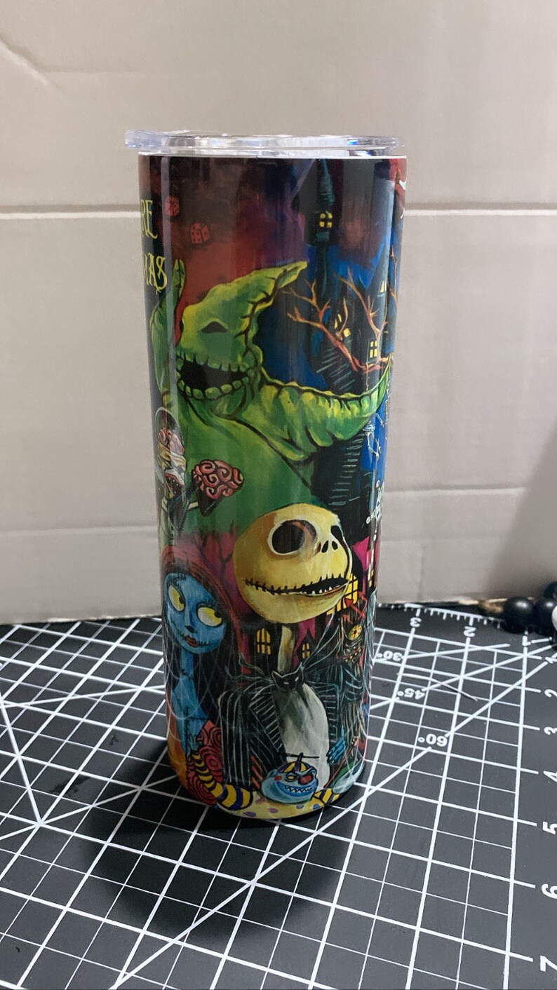 K&ouml;nnte beinhalten: Ein Edelstahlbecher mit Figuren aus "The Nightmare Before Christmas". Das Design umfasst Jack Skellington, Sally, Oogie Boogie und andere Charaktere vor einem dunklen, gruseligen Hintergrund. Der Becher hat einen transparenten Deckel.