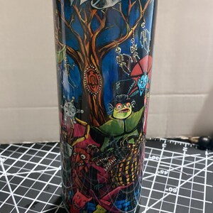K&ouml;nnte beinhalten: Ein hoher, zylindrischer Becher mit einer farbenfrohen, detaillierten Illustration von Figuren und Szenen aus einem Fantasy-Film. Das Design umfasst einen Baum, Skelette und andere gruselige Figuren vor einem dunkelblauen Hintergrund.