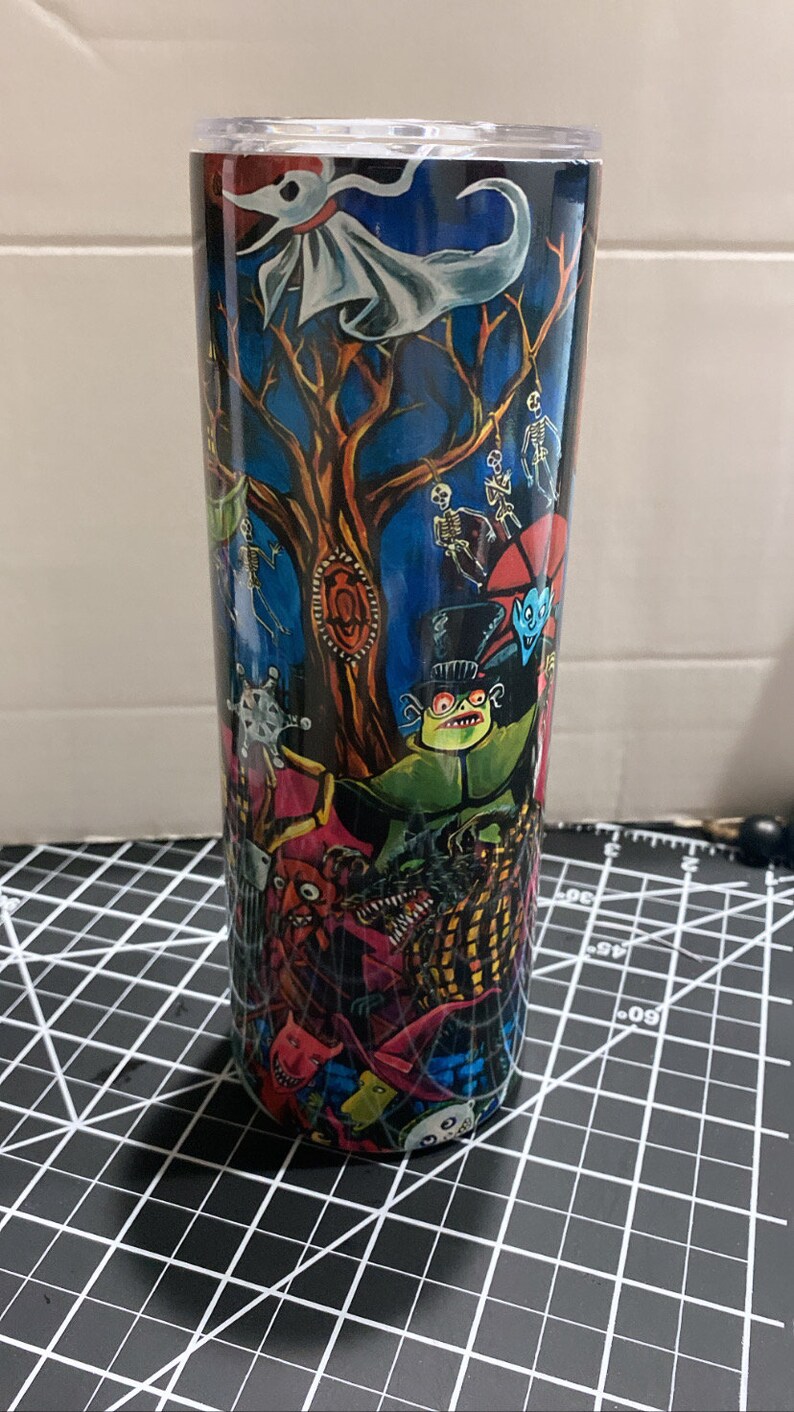 K&ouml;nnte beinhalten: Ein hoher, zylindrischer Becher mit einer farbenfrohen, detaillierten Illustration von Figuren und Szenen aus einem Fantasy-Film. Das Design umfasst einen Baum, Skelette und andere gruselige Figuren vor einem dunkelblauen Hintergrund.