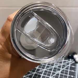 K&ouml;nnte beinhalten: Nahaufnahme eines Deckels f&uuml;r einen Edelstahlbecher mit einem transparenten Schiebeverschluss. Der Deckel ist transparent und gibt den Blick auf das Innere des Bechers frei. Er hat einen wei&szlig;en, ovalen Schieber und einen kleinen, wei&szlig;en Verschluss.