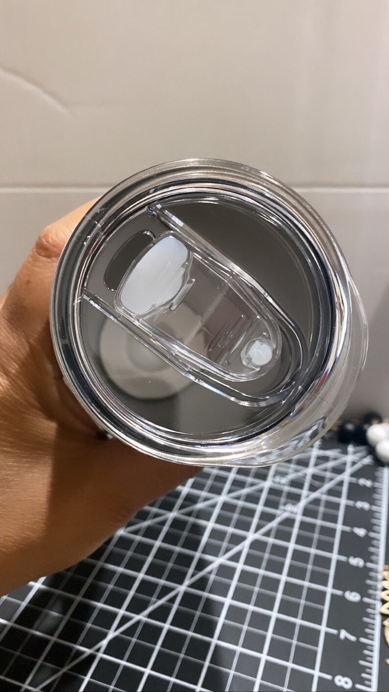 K&ouml;nnte beinhalten: Nahaufnahme eines Deckels f&uuml;r einen Edelstahlbecher mit einem transparenten Schiebeverschluss. Der Deckel ist transparent und gibt den Blick auf das Innere des Bechers frei. Er hat einen wei&szlig;en, ovalen Schieber und einen kleinen, wei&szlig;en Verschluss.