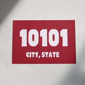 Può includere: Un cartello rettangolare rosso con testo bianco. Il numero 10101 è stampato in grande carattere bianco. Sotto il numero c'è il testo "CITY, STATE" in carattere bianco più piccolo.