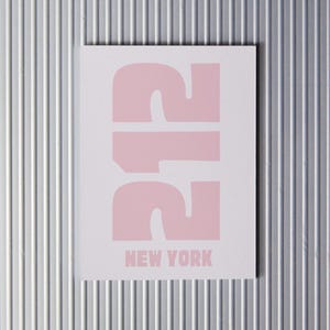 Puede incluir: Una tarjeta blanca con el número "212" impreso en rosa, en fuente grande y negrita. El texto "NEW YORK" está impreso en negro debajo del número.