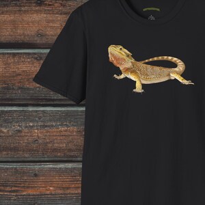 Op de afbeelding: Zwart T-shirt met een afbeelding van een baardagame hagedis. De hagedis is bruin en geel met een stekelige kam op zijn kop. De tekst "Creepy Crawly Apparel" staat in de rechterbovenhoek van de afbeelding.