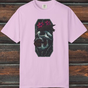 May include: Light pink t-shirt with a graphic of a skull, raven, and flowers. The graphic is in a dark, rectangular frame. The text "Creepy Crawly Apparel" is in the upper right corner.