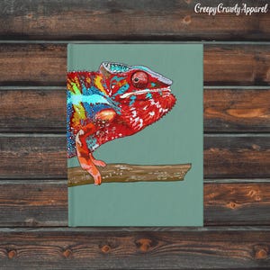 Puede incluir: Ilustración de un camaleón colorido sobre un fondo verde azulado, posado en una rama marrón. El reptil presenta tonos rojos, azules y amarillos. El texto "Creepy Crawly Apparel" está en la esquina superior derecha.