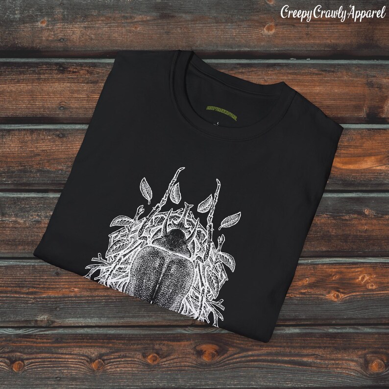 Op de afbeelding: Zwart T-shirt met een witte grafische print van een kever omringd door bladeren. De tekst "Creepy Crawly Apparel" is in wit bovenaan de afbeelding gedrukt.