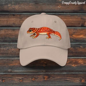 May include: A beige baseball cap featuring a detailed illustration of an orange and white spotted gecko centred on the front panel. The gecko is depicted in profile, facing left, with large dark eyes and a textured, patterned body design.