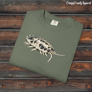 May include: An olive green t-shirt featuring a detailed illustration of a woodlouse. The woodlouse is depicted in shades of black, white, and tan, with multiple legs and antennae. The text "Creepy Crawly Apparel" is in the top right corner.