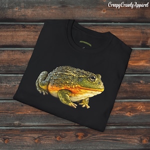 May include: Black t-shirt featuring a detailed illustration of a frog with green, yellow, and orange tones. The frog is the focal point, set against a dark background. The text "Creepy Crawly Apparel" is in the upper right corner.
