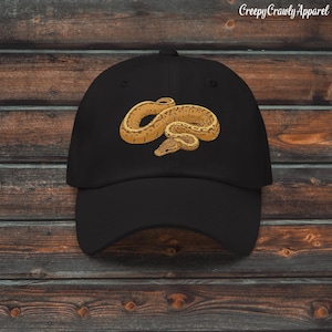 Può includere: Cappellino da baseball nero con un'illustrazione dettagliata di un serpente nei toni del marrone e del beige. Il testo "Creepy Crawly Apparel" è stampato in bianco in alto a destra sul cappellino.