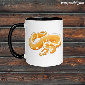 Puede incluir: Taza de cerámica blanca con asa y borde negros. La taza presenta una ilustración detallada de una serpiente naranja y blanca. El texto "Creepy Crawly Apparel" está impreso en la parte superior derecha de la taza.