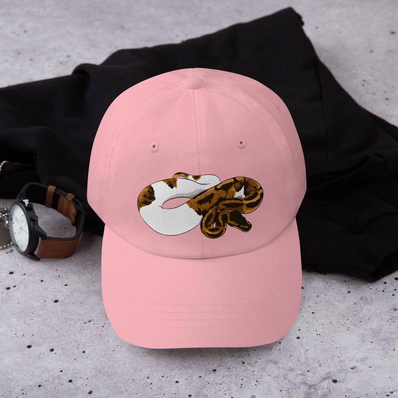 May include: A light pink baseball cap featuring a graphic of a snake in shades of brown, white, and black. The cap is made of a soft material and has a curved brim. A black jacket and a watch with a brown strap are in the background.