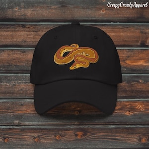 Può includere: Cappellino da baseball nero con un disegno di serpente ricamato nei toni del marrone, giallo e rosso. Il cappellino ha una visiera curva e una classica costruzione a sei pannelli. Il testo "Creepy Crawly Apparel" è stampato in alto a destra.