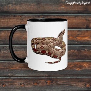 Pode incluir: Caneca de cerâmica branca com alça e borda pretas, com uma ilustração detalhada de uma cobra marrom e vermelha. A caneca tem o texto "Creepy Crawly Apparel" impresso no canto superior direito.