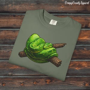 Puede incluir: Camiseta verde oliva con una ilustración detallada de una serpiente verde enrollada en una rama marrón. La serpiente tiene ojos amarillos y una textura escamosa. El texto "Creepy Crawly Apparel" está en la esquina superior derecha.