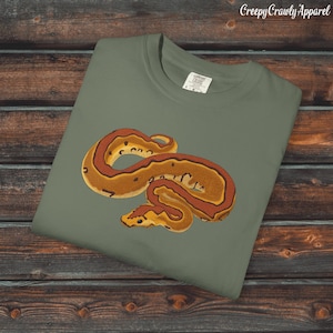 May include: A sage green t-shirt with a detailed illustration of a snake in brown, orange, and yellow. The snake is coiled and centered on the shirt. The text "Creepy Crawly Apparel" is in the upper right corner.