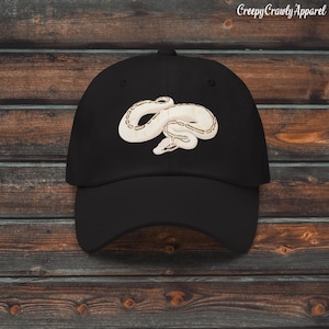 Könnte beinhalten: Schwarze Baseballkappe mit einem cremefarbenen Schlangenmuster. Die Schlange ist gewunden und mit braunen Akzenten versehen. Der Text "Creepy Crawly Apparel" ist oben rechts im Bild aufgedruckt.