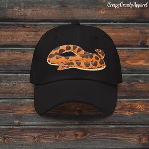 Pode incluir: Boné de beisebol preto com um desenho de cobra de desenho animado em tons de laranja e castanho. O boné tem uma aba curva e uma construção clássica de seis painéis. O texto "Creepy Crawly Apparel" está impresso no canto superior direito.