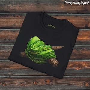 Może przedstawiać: Czarny t-shirt z szczegółową ilustracją jasnozielonego węża owiniętego wokół brązowej gałęzi. Łuski węża są teksturowane. Tekst "Creepy Crawly Apparel" jest widoczny w prawym górnym rogu.